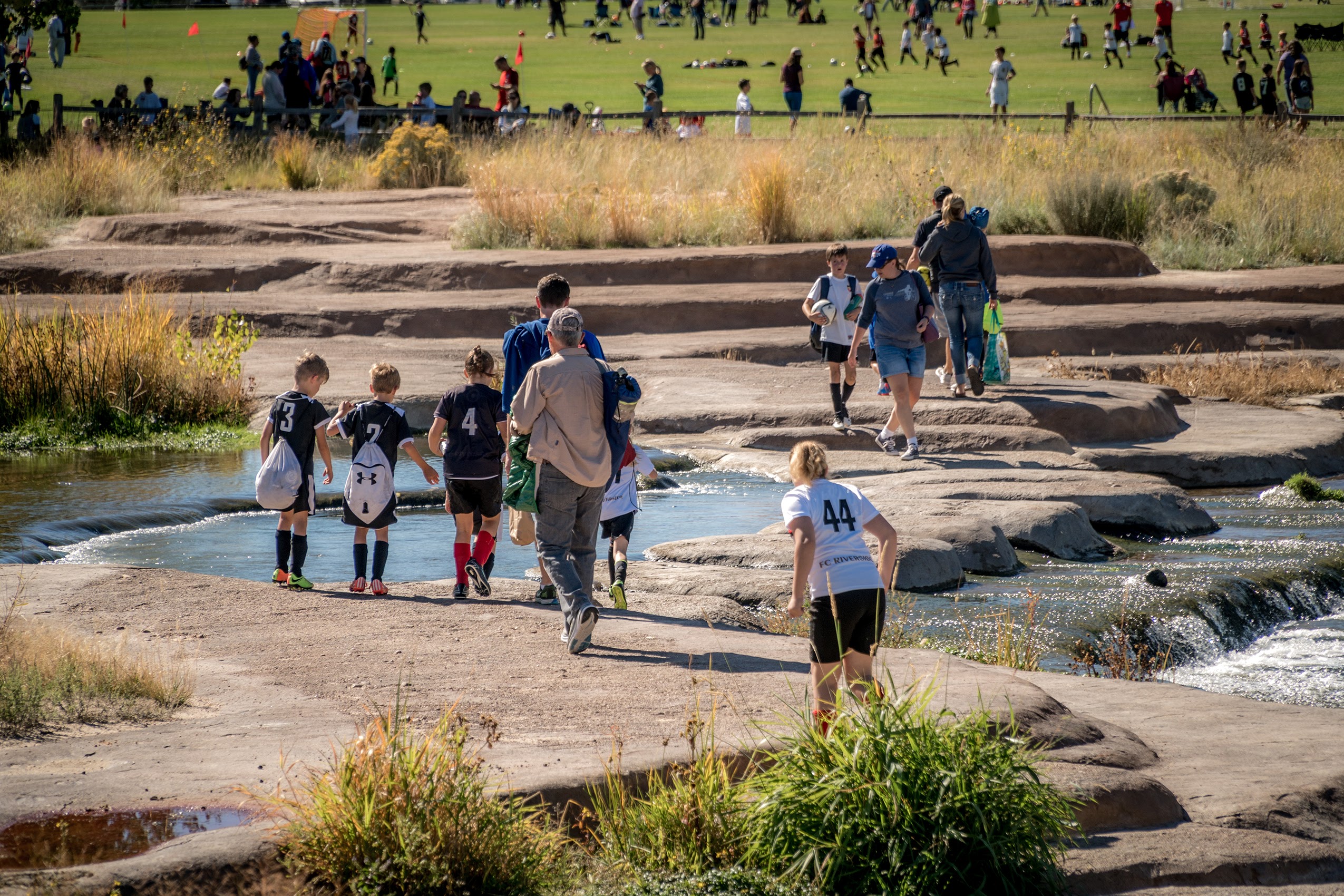 Adults and children cross an environmental stepping stone feature over a river